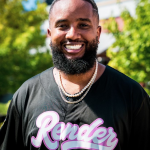 Headshot photograph of Justin Samuels, a Black man who is wearing two chain necklaces and a jersey that says Render in pink script.