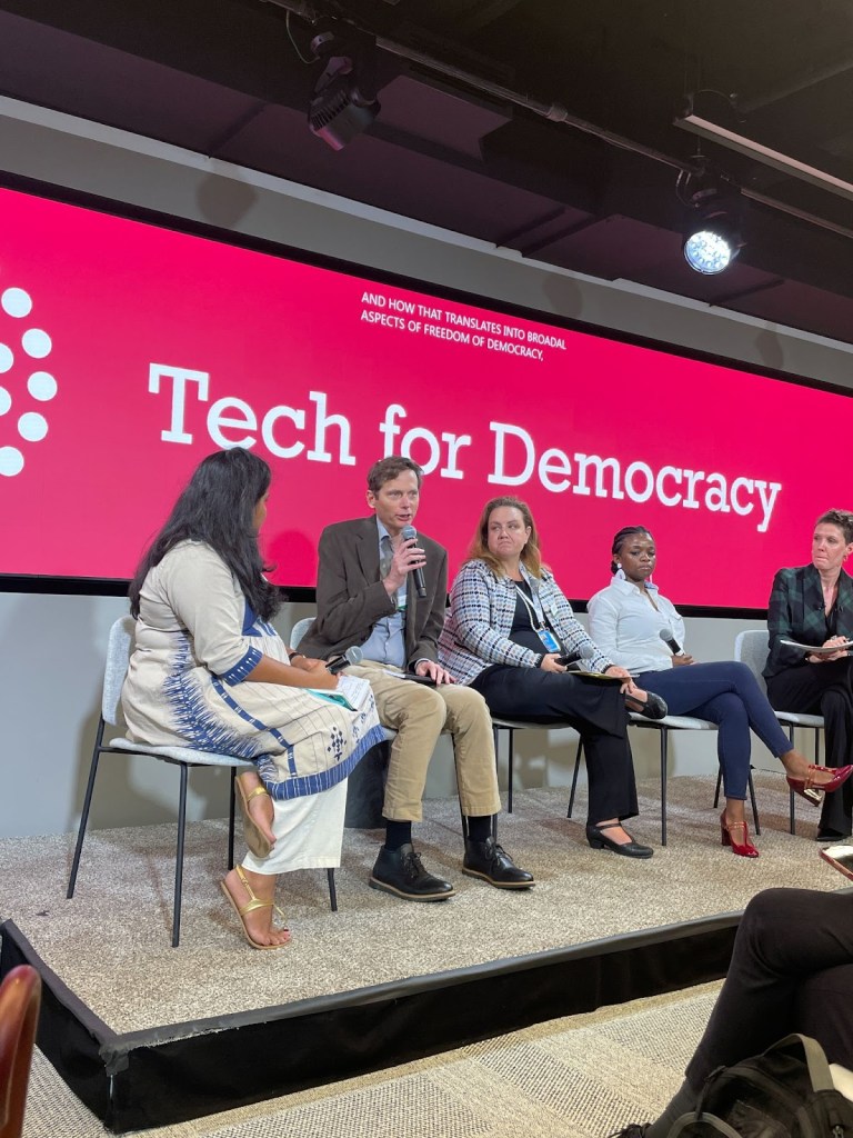 The five panelists on the  the Tech for Democracy high-level side-event at UNGA77 sitting in front of a pink screent hat reads "Tech for Democracy." Mike Linksvayer, Head of Policy at GitHub, is speaking into a handheld microphone.
