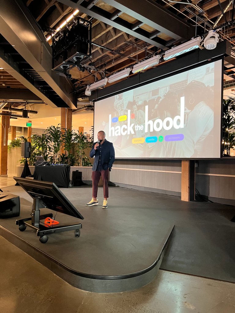 A presenter stands in front of a screen that reads "Hack the Hood" on the stage in GitHub HQ in San Francisco.