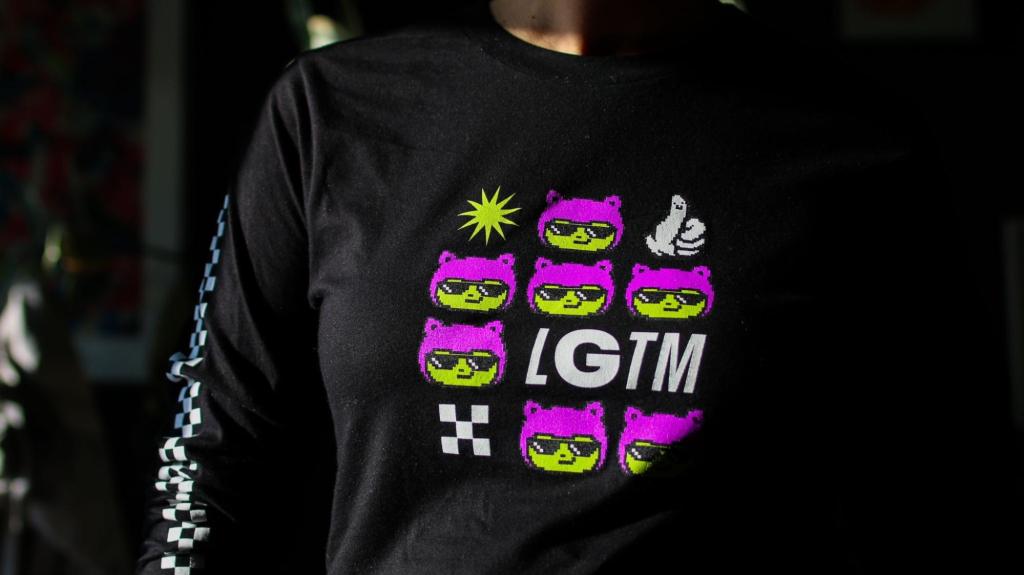A close-up of the design on a black long-sleeved t-shirt. The design includes green and purple Octocats, a thumbs up emoji, and the letters 