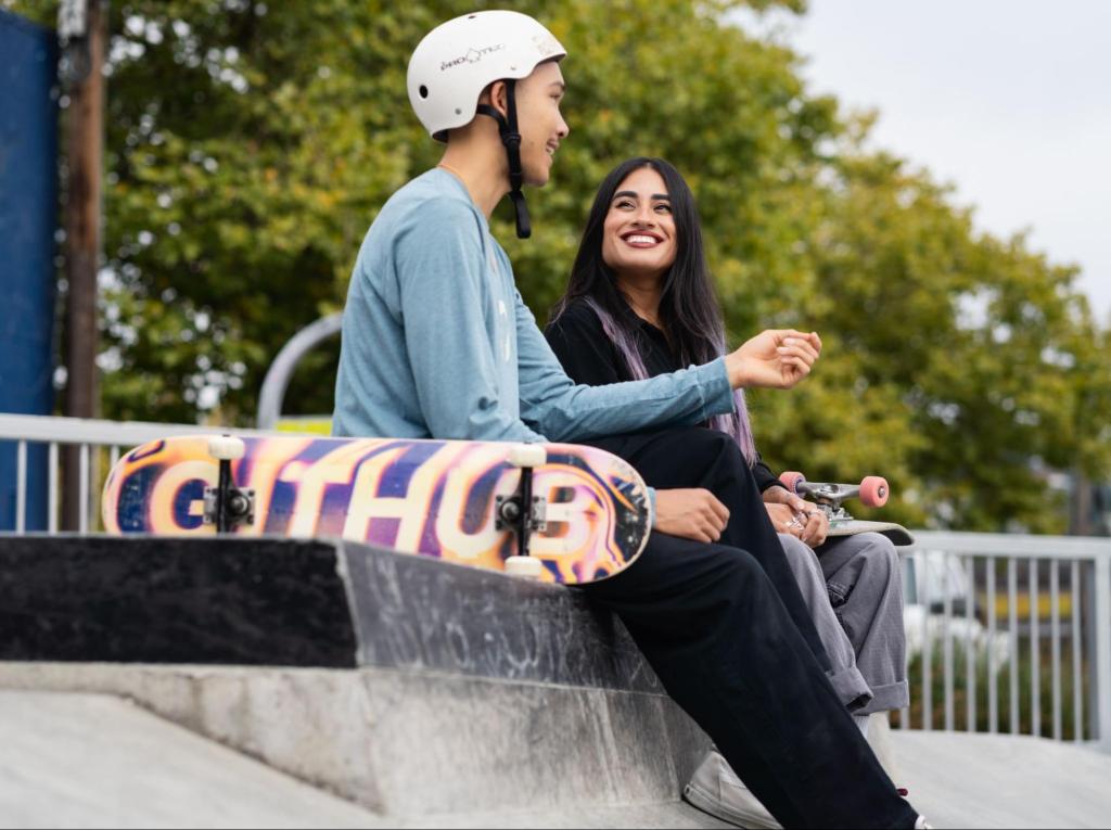 Two smiling people are sitting on the edge of a skatepark bowl. One is wearing a white helmet and has his skateboard perched next to him, with the GitHub-branded deck visible to the camera.