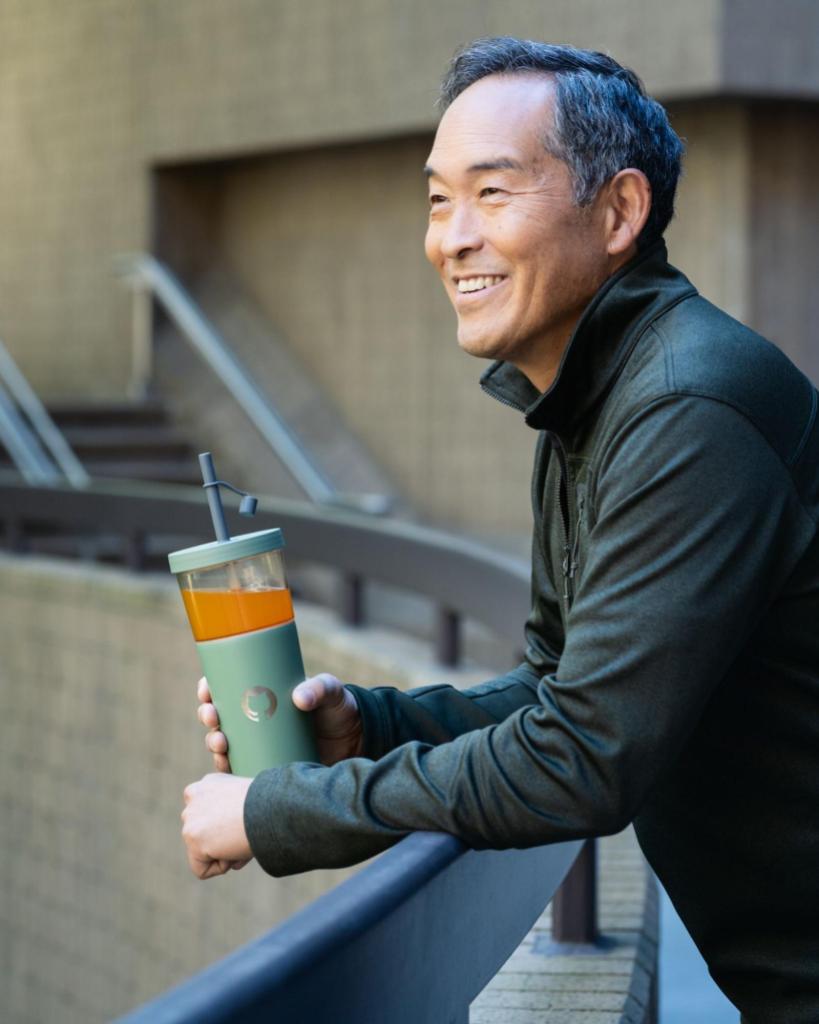 A smiling man leans against the railing of a balcony holding a green GitHub-branded tumbler filled with an orange drink.