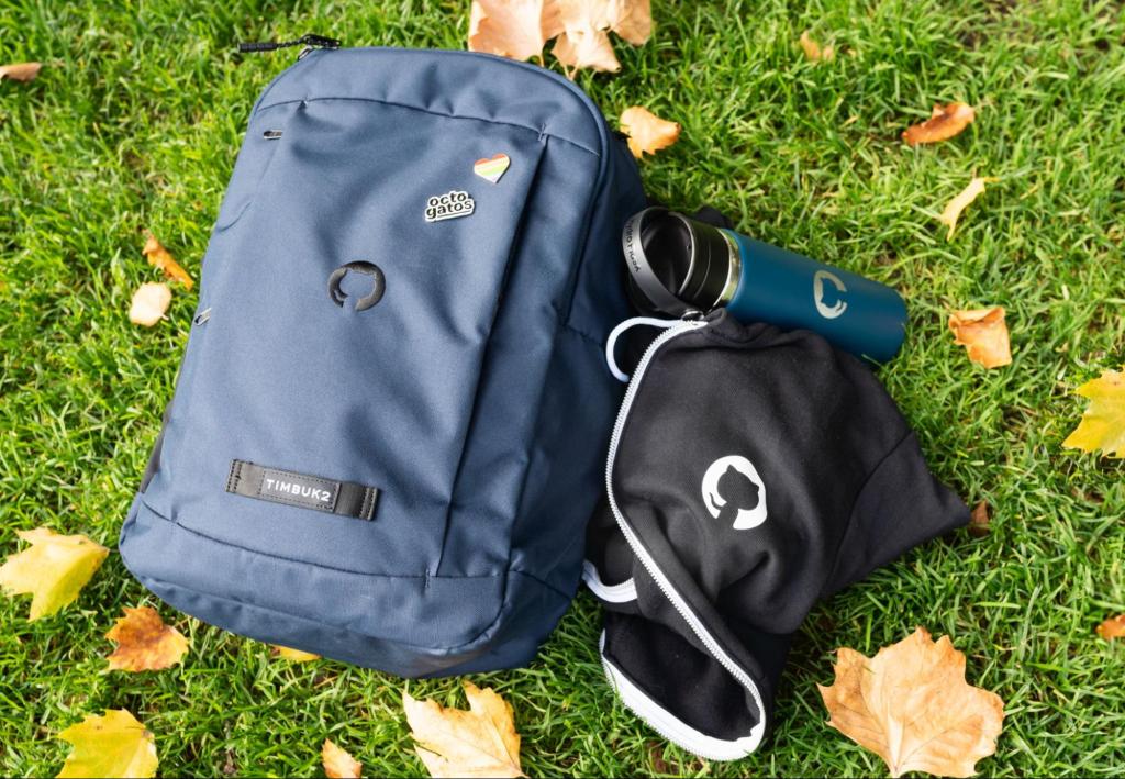 A GitHub-branded backpack, an Octocat logo hoodie, and an insulated tumbler with an octocat logo sit in a pile on some bright green grass.