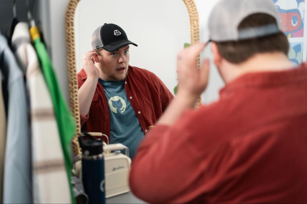 A man wearing a GitHub-branded trucker style cap looks into a mirror and fixes his hair.