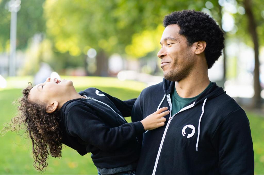 A man smiles at a small girl who he is holding in his arms. Both are wearing black hoodies with the Octocat logo on them.