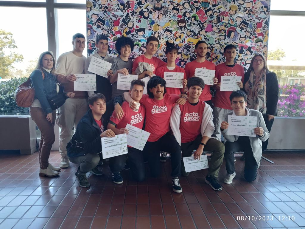 A group of students wearing red t-shirts that read "Octogatos." They are holding certificates and standing in front of a large banner with a collage of GitHub stickers on it.