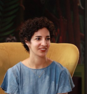 A photograph of Nadine Krish Spencer, a woman with short, dark curly hair who is smiling and sitting in a yellow wingback armchair.