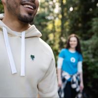 A photo of someone wearing the GitHub Sportiqe hoodie with a green invertocat patch. In the background is someone wearing the fitted invertocat blue tee.