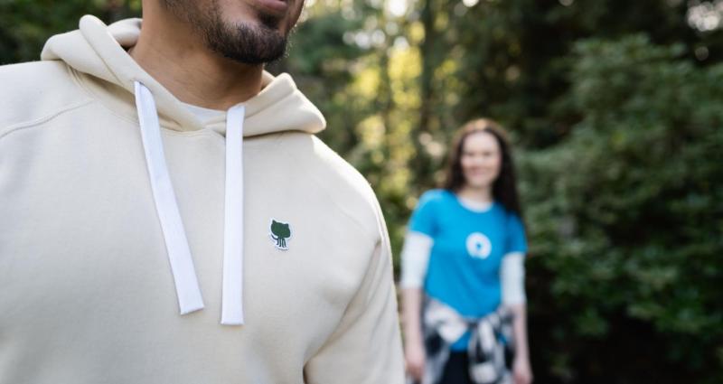 一张某人穿着GitHub Sportiqe连帽衫和绿色内底贴片的照片。背景中有人穿着合身的蓝色T恤。