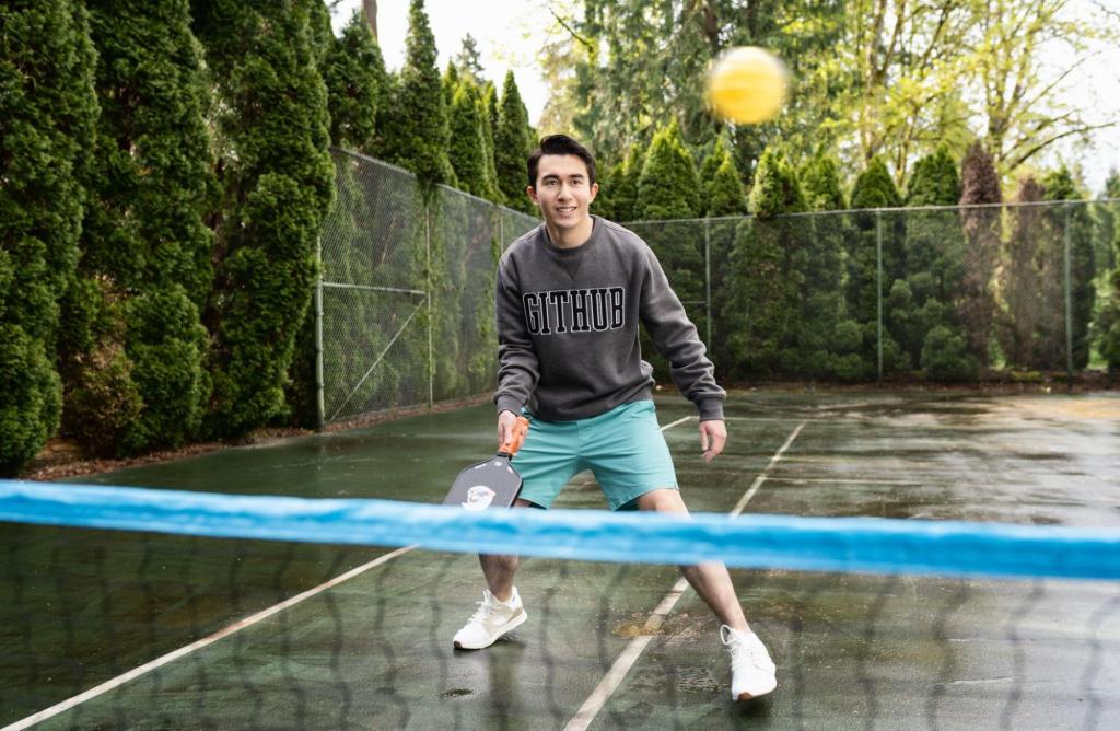 A smiling man wielding a pickleball paddle is wearing a grey sweatshirt with 