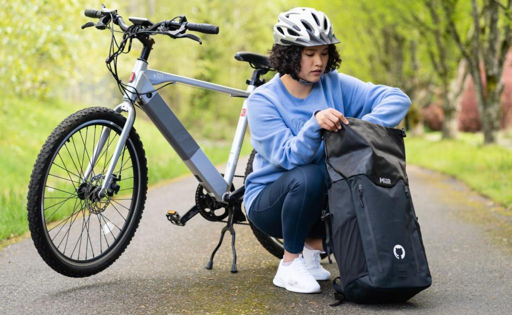 A woman wearing a bike helmet kneels down next to her bike to reach into a black Miir brand backpack with a white octocat logo on it. Her blue crew neck sweatshirt also says GitHub on it.