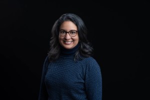 Headshot photograph of Seema Iyer, senior director of The Hive at USA for UNHCR. She is smiling at the camera and has dark shoulder-length hair, wearing dark framed glasses and a dark blue turtleneck sweater.