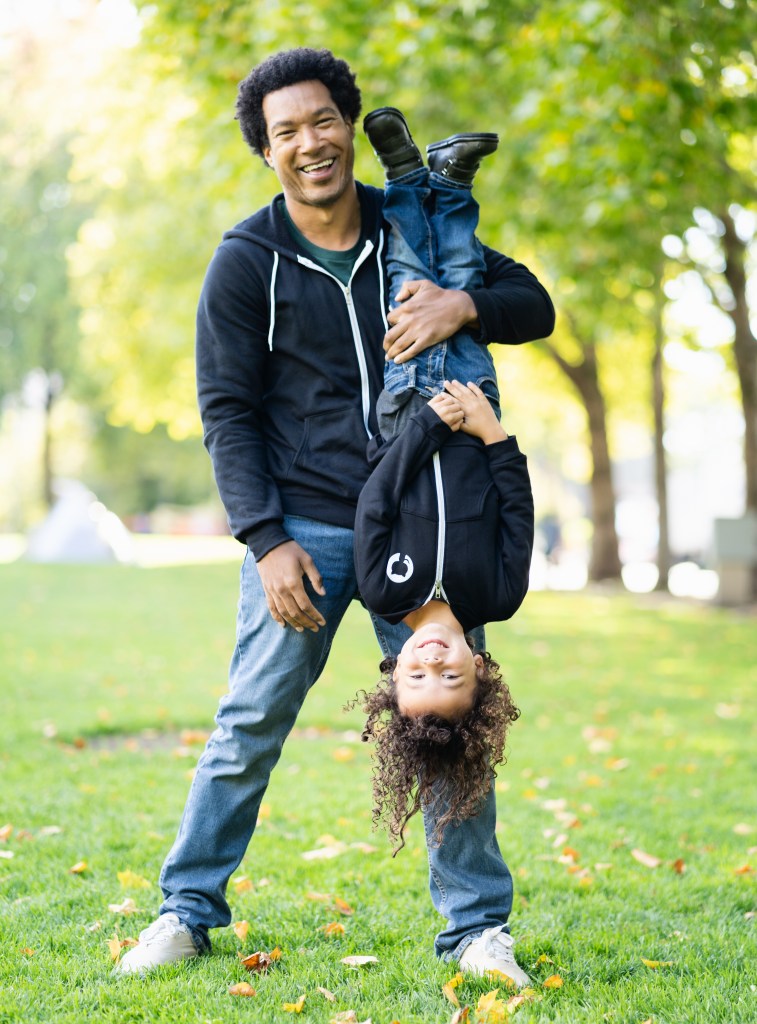 Man wearing a black GitHub hoodie holding a kid upside down who is also wearing a black GitHub hoodie.