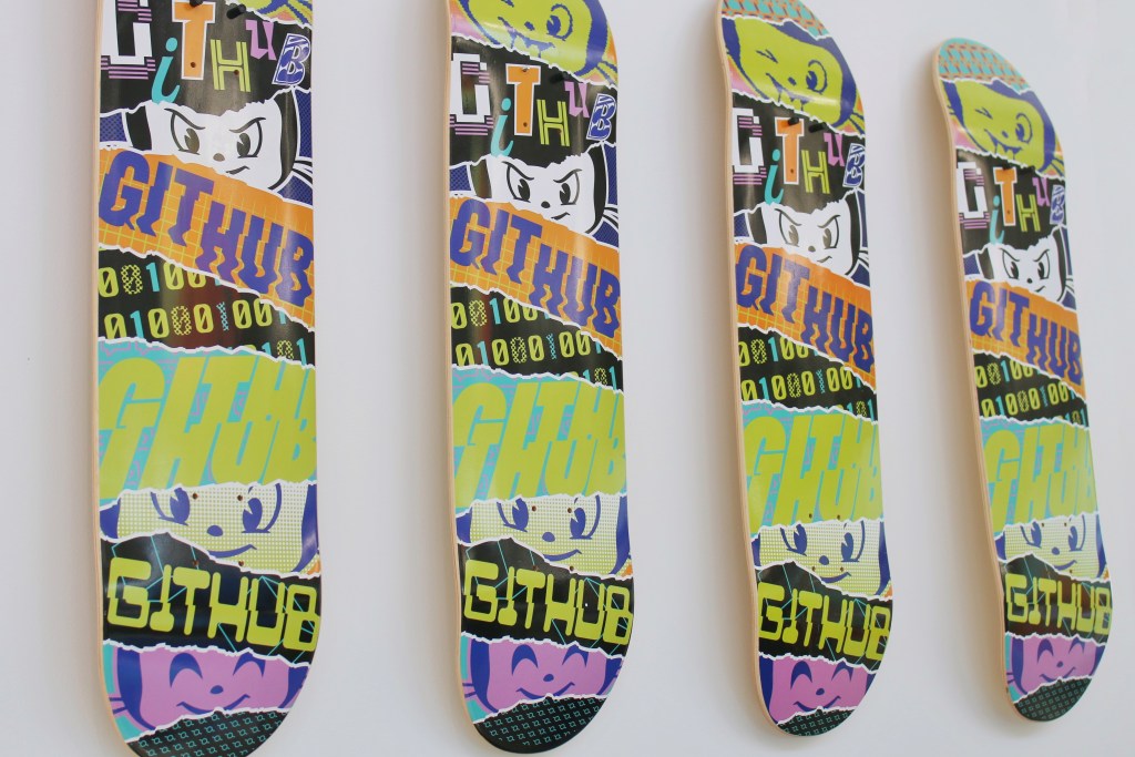 Four skateboard decks displayed in a row against a white background, featuring colorful and layered graphics with bold text "GITHUB" in multiple orientations. The designs include cartoon-like characters, binary numbers, and vibrant colors like yellow, orange, purple, and green.