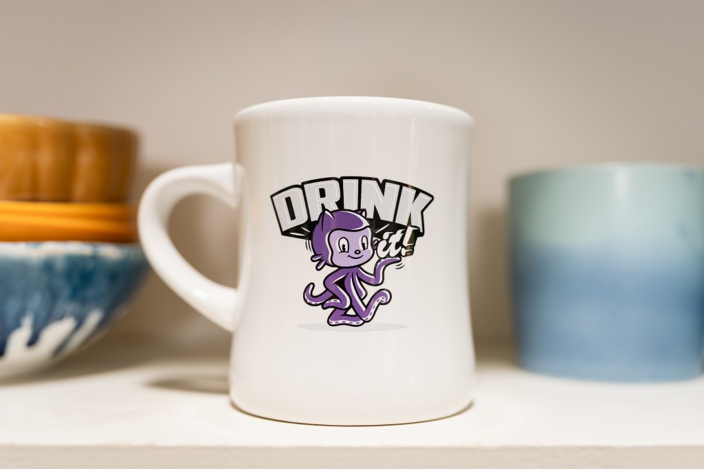 Close up of the GitHub Diner Mug with an Octocat and text says Drink it! In the background are other cups and bowls.