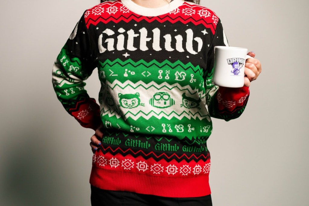 Person wearing a red, black and green ugly sweater with GitHub designs and Octocats. They are holding a GitHub Diner Mug with an Octocat with the text Drink it.