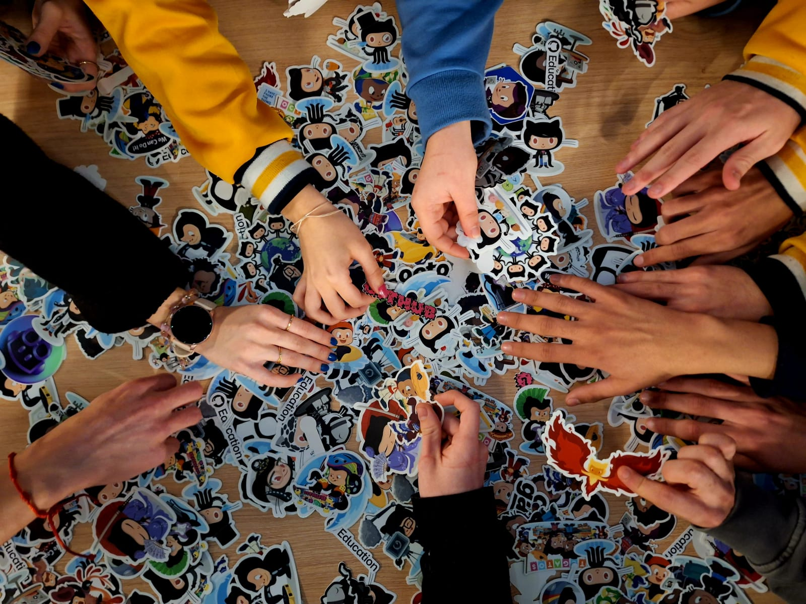 Several people's hands are reaching into a big pile of stickers on top of a table.