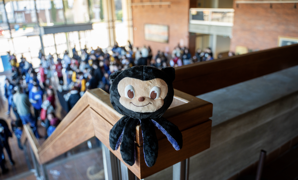 A Mona the octocat plushie perched on a stair railing, with a large group of people softly visible in the background.