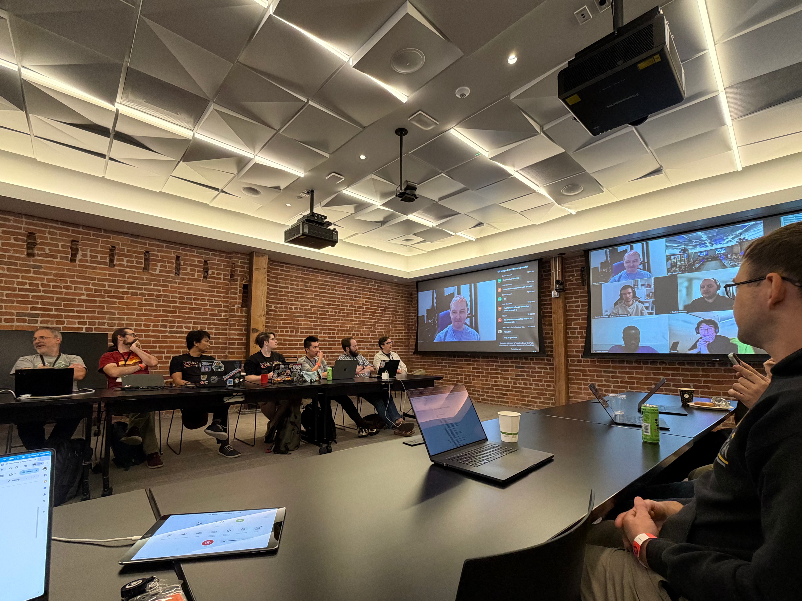 People seated around a long conference table at GitHub HQ’s Executive Briefing Center during the Git Contributors Summit. Laptops are open, and several participants join remotely on large screens at the front of the room.