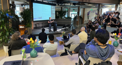 GitHub’s PJ Metz speaks on stage in the GitHub HQ auditorium, addressing a large audience seated on tiered benches. The space features exposed beams, plants, and a hanging green Invertocat installation overhead.