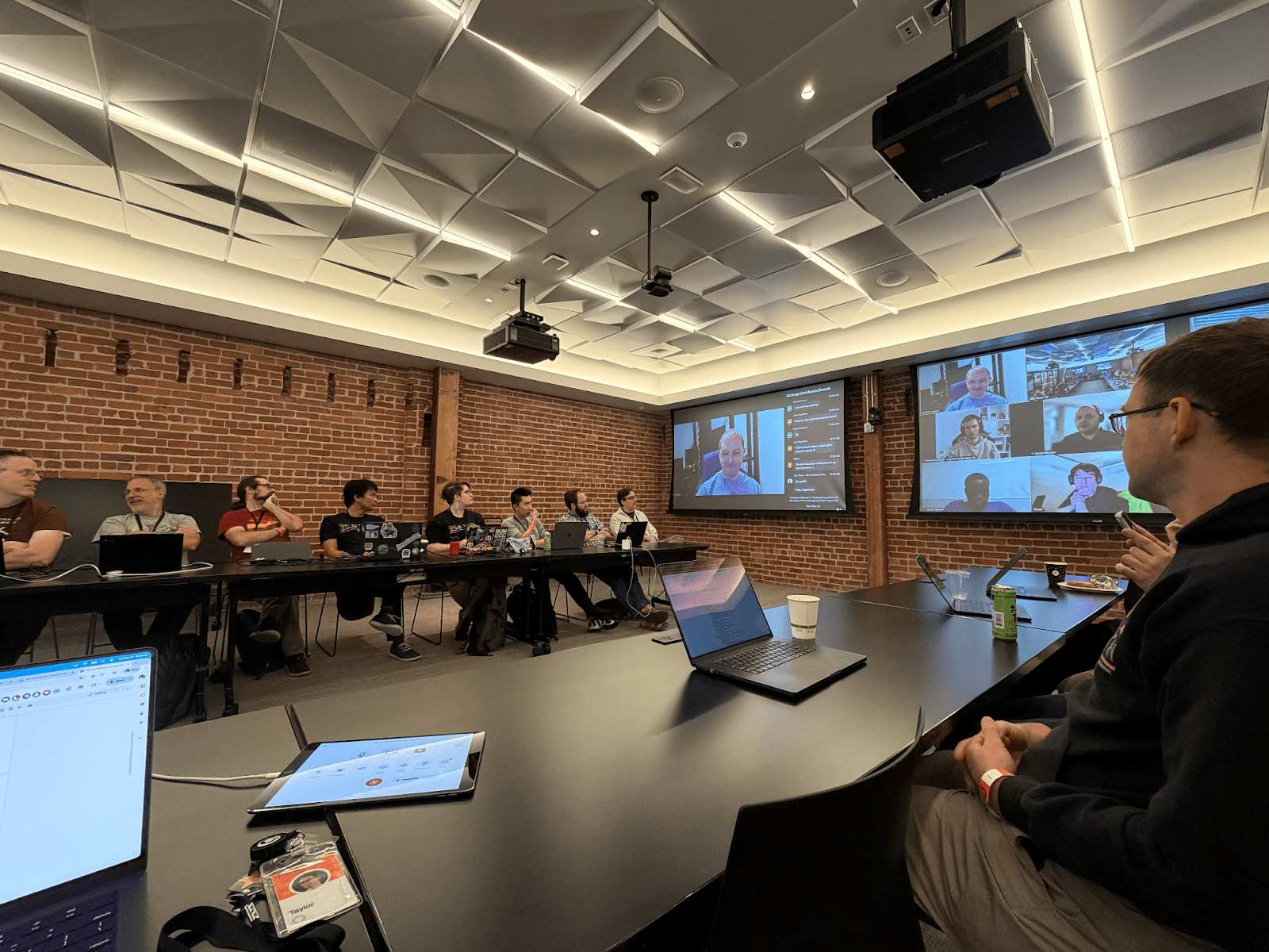 People seated around a long conference table at GitHub HQ’s Executive Briefing Center during the Git Contributors Summit. Laptops are open, and several participants join remotely on large screens at the front of the room.