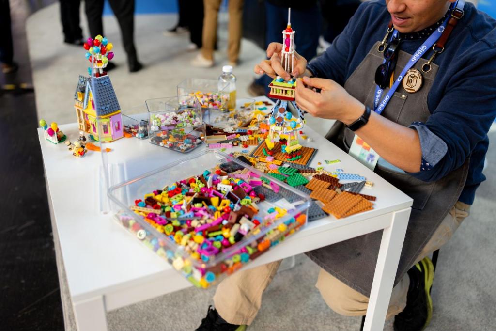 Person at table assembling colorful LEGO buildings including a tower and house with organized brick containers.